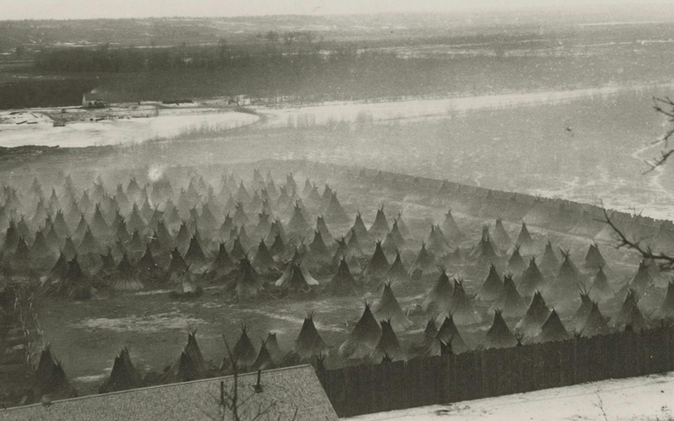 A photograph shows dozens of tepees fenced in by a tall stockade. The ground appears to be frozen mud. Smoke rises from several tepees, the result of warming fires within